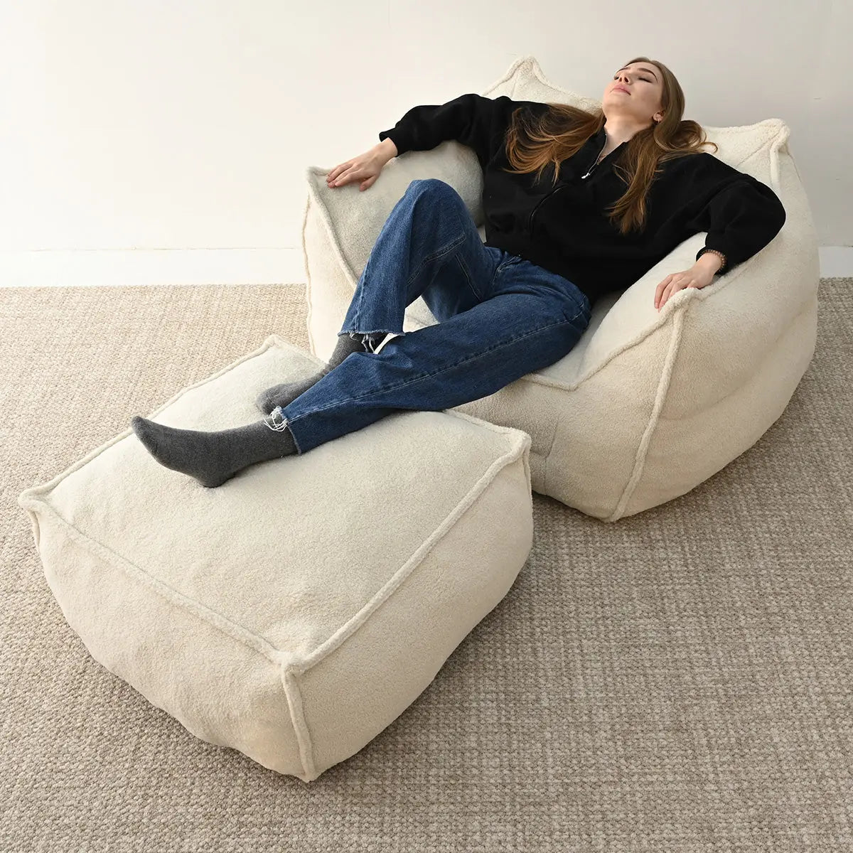 Person relaxing on beige Boring Teddy Bean Bag Chair & Ottoman Set, wool rug, minimalist room.