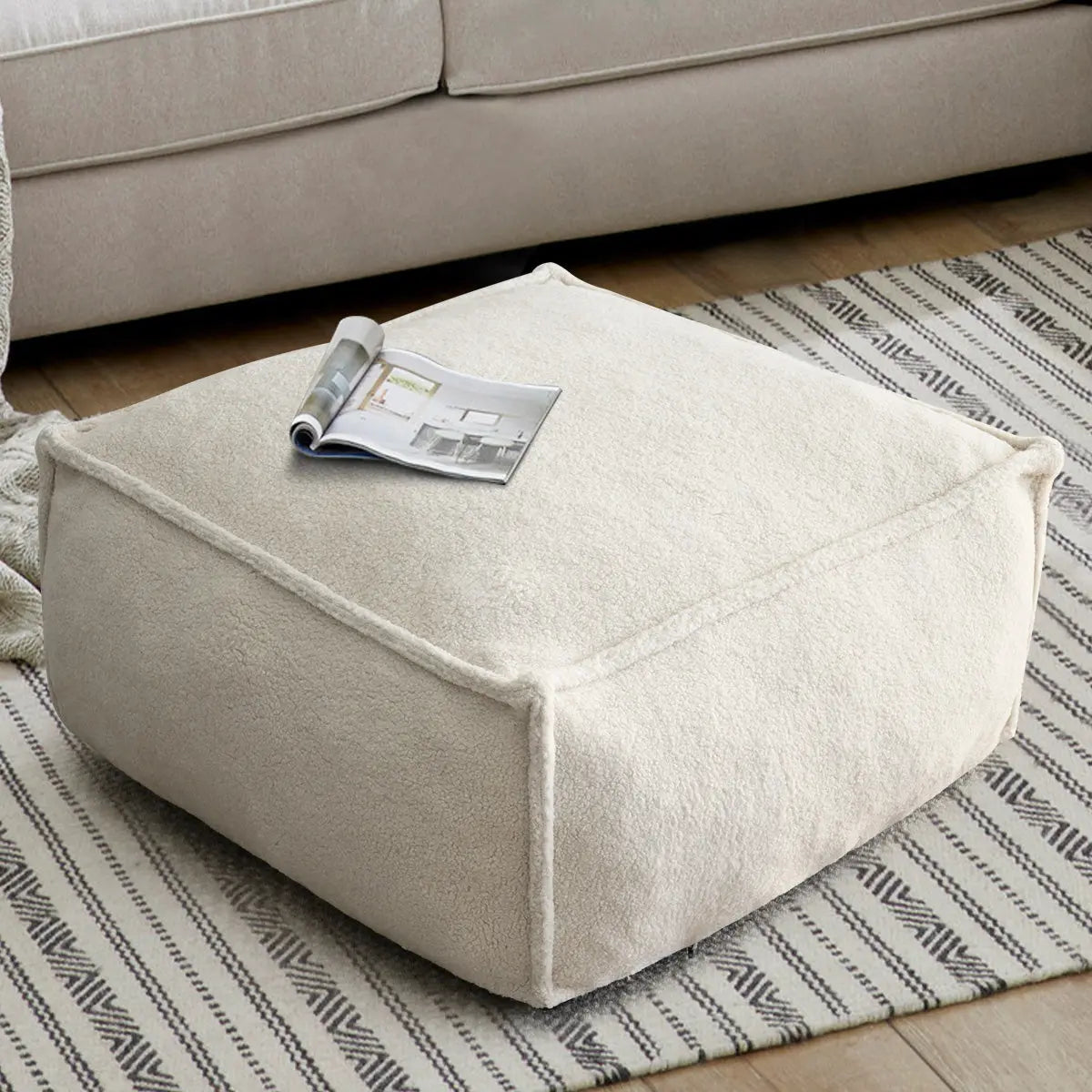 Boring Teddy square bean bag ottoman on patterned rug, next to beige sofa, wooden flooring.