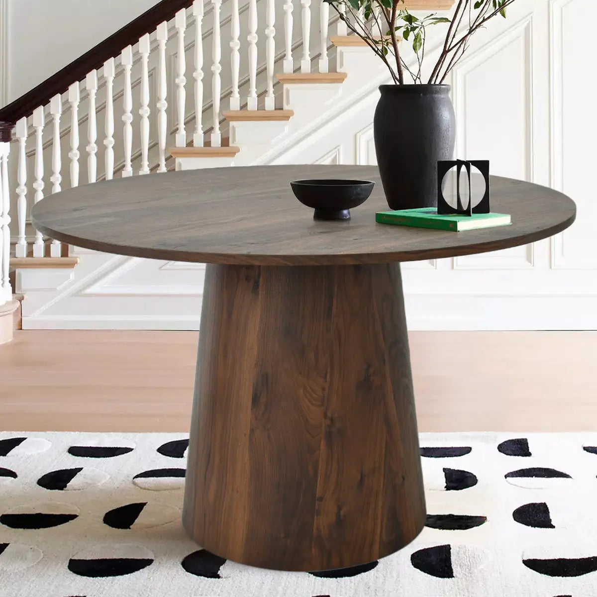 Dwen Modern Round Pedestal Dining Table, wooden finish, staircase backdrop, patterned rug, beige flooring.