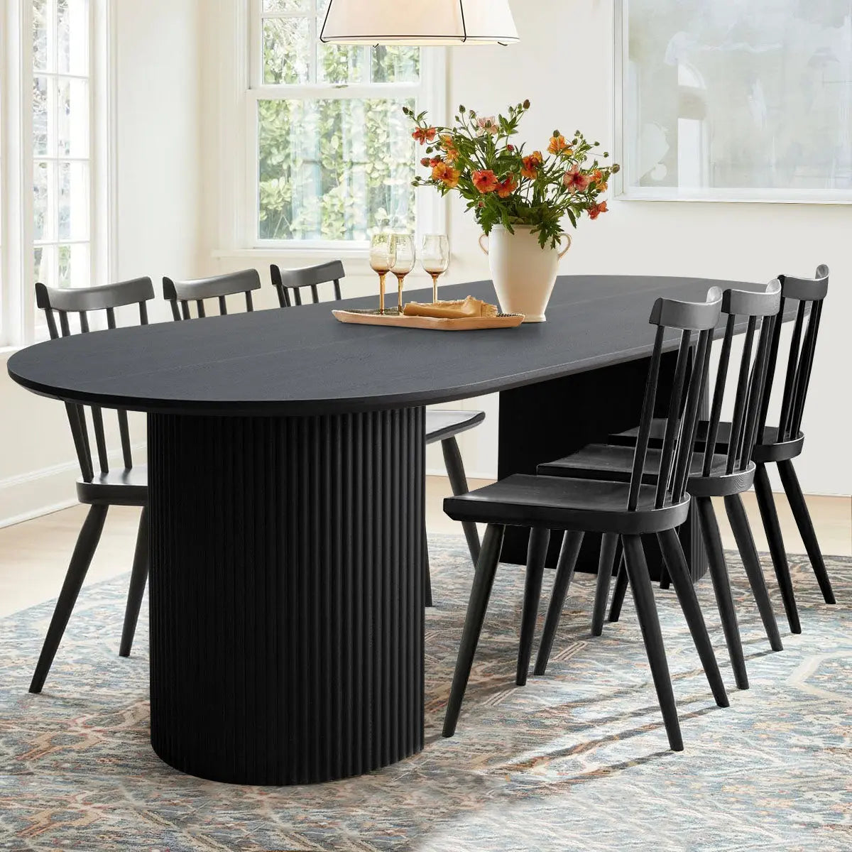 Dwen Modern Large Oval Dining Table with Roman pillar base, black chairs, patterned rug, bright room.