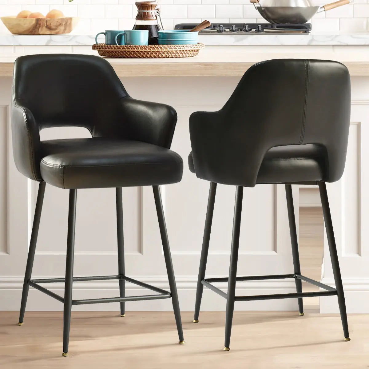 Black faux leather counter stools with arms, kitchen setting, light wood flooring, white panel walls.