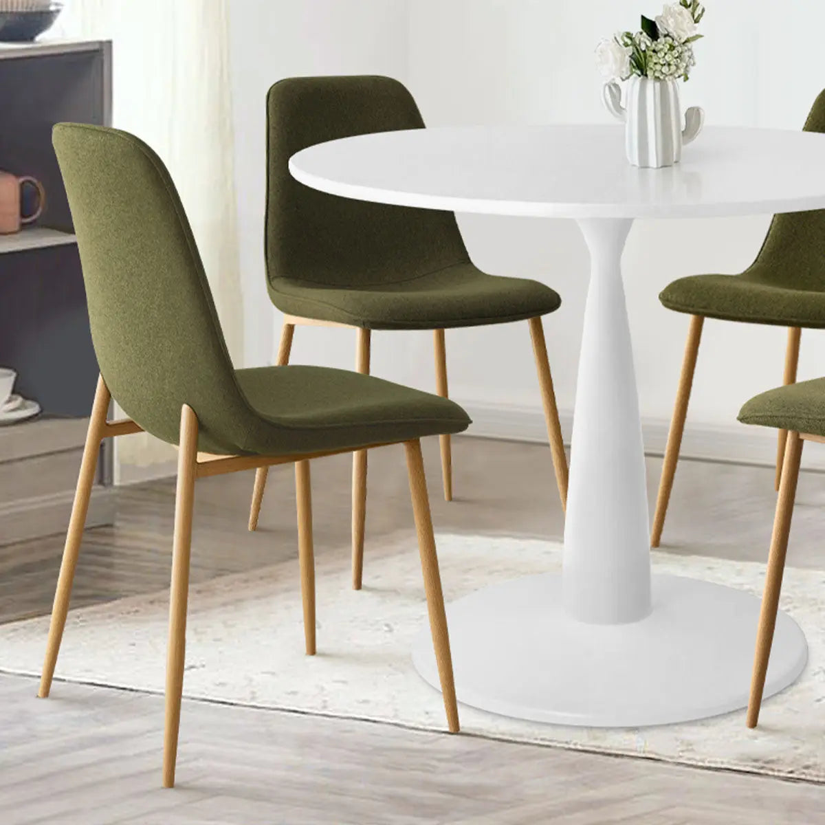 Haven & Oslo white round dining table set with olive chairs, light wood legs, white wall, wood floor.