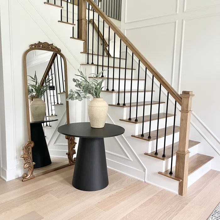 Dwen 35" round pedestal console table by staircase, wood flooring, ornate mirror, light walls.