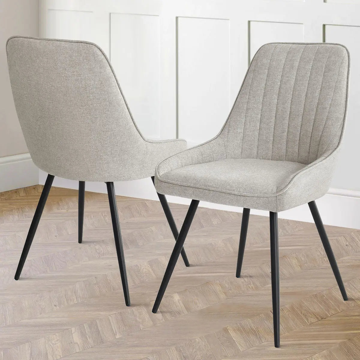 Boston Modern Upholstered Dining Chair, light gray fabric, black legs, wooden herringbone floor, white paneled wall.