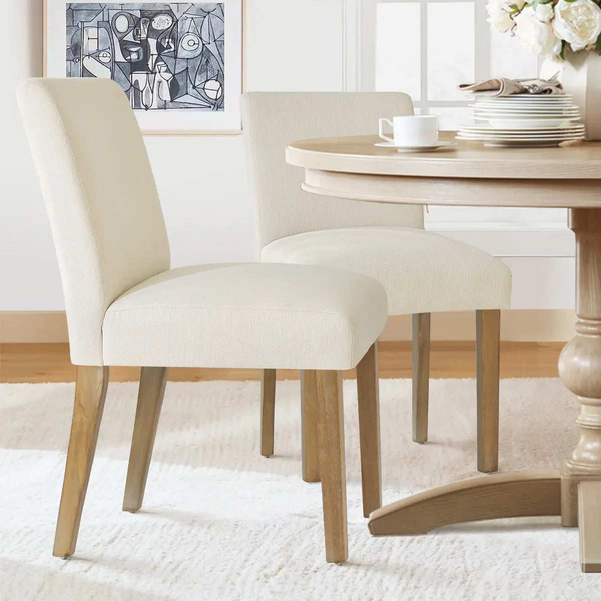 Beige North Upholstered Dining Chair with light wood legs, round wooden table, white rug, abstract artwork.